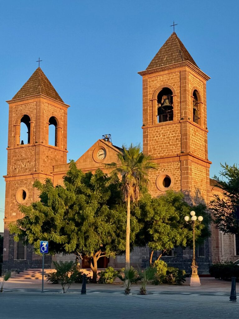 Zwei steinerne Kirchtürme mit spitzen Dächern und Kreuzen, davor Bäume und eine Straßenlaterne. Es handelt sich um die Catedral de Nuestra Señora de La Paz in Baja California Sur