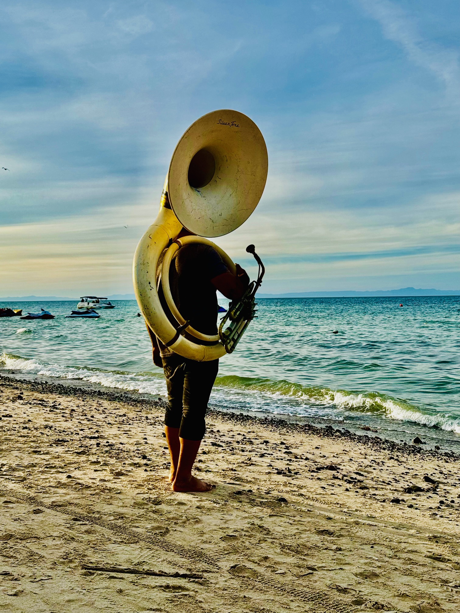 Person steht barfuß am Strand und spielt eine große, goldfarbene Sousaphon-Tuba, im Hintergrund Meer und bewölkter Himmel