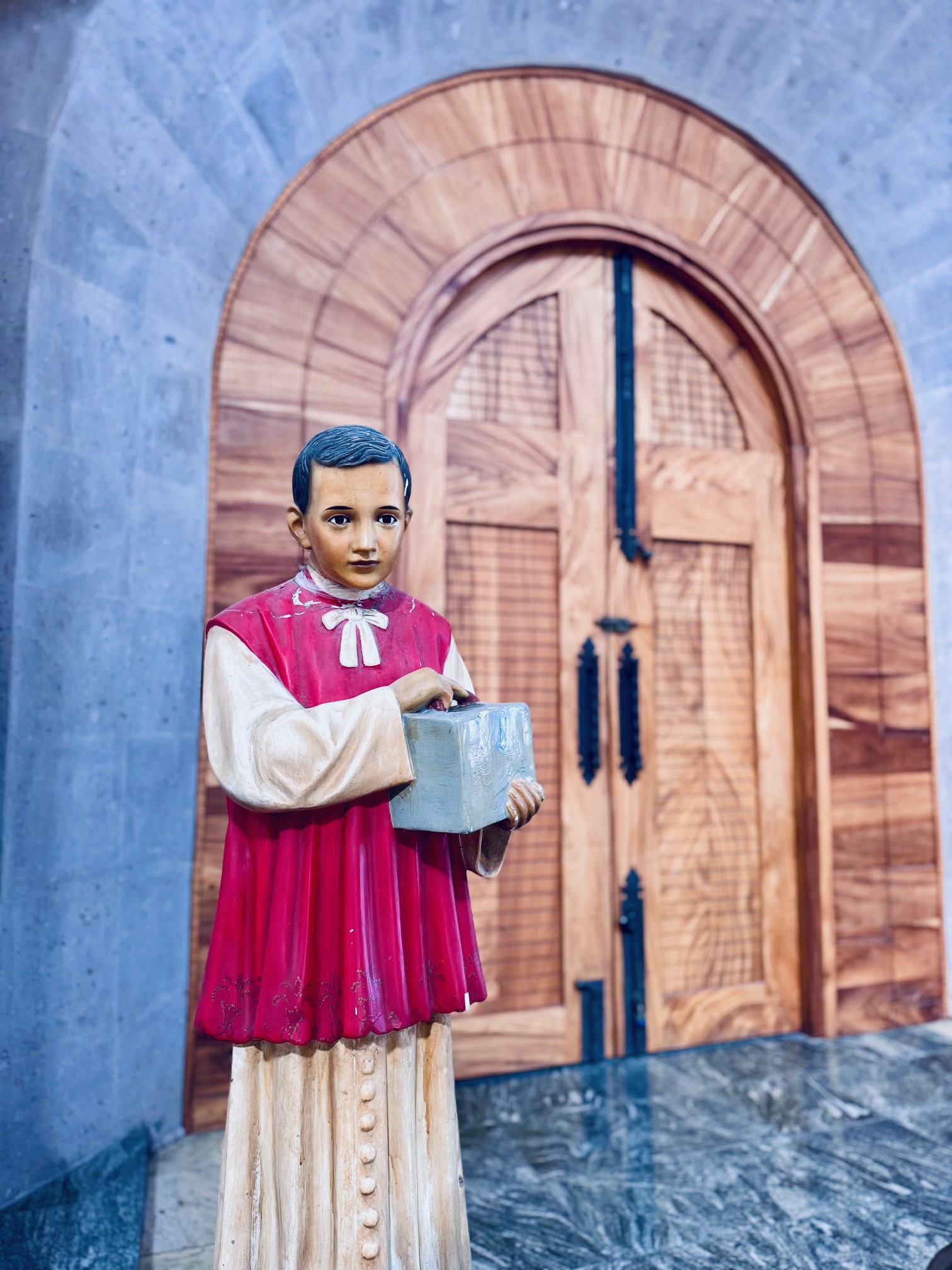 Opferstock in Form eines Jungen in rotem Messdienergewand mit weißem Kragen, der eine graue Schachtel hält, vor einer großen hölzernen Bogentür