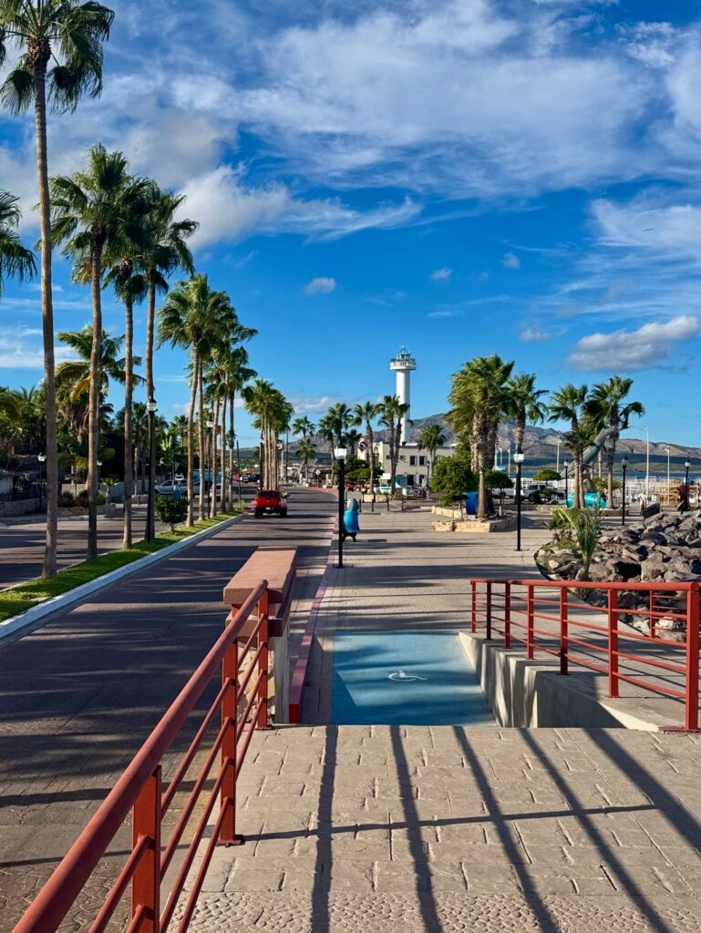 Promenade mit Palmen, rotem Geländer und weißem Leuchtturm im Hintergrund unter blauem Himmel mit freundlichen Wolken