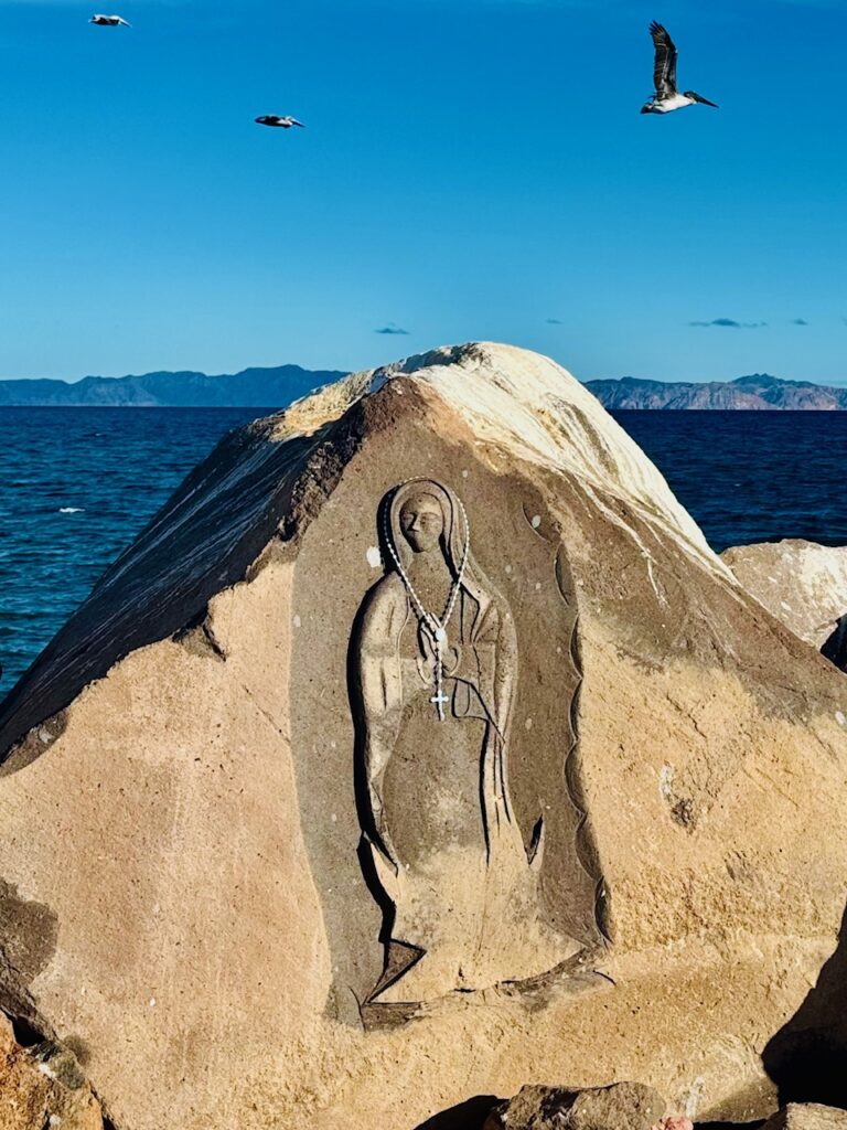 Fels mit eingemeißelter Figur der Jungfrau Maria, die einen echten Rosenkranz trägt. Im Hintergrund Meer und zwei Möwen vor blauem Himmel