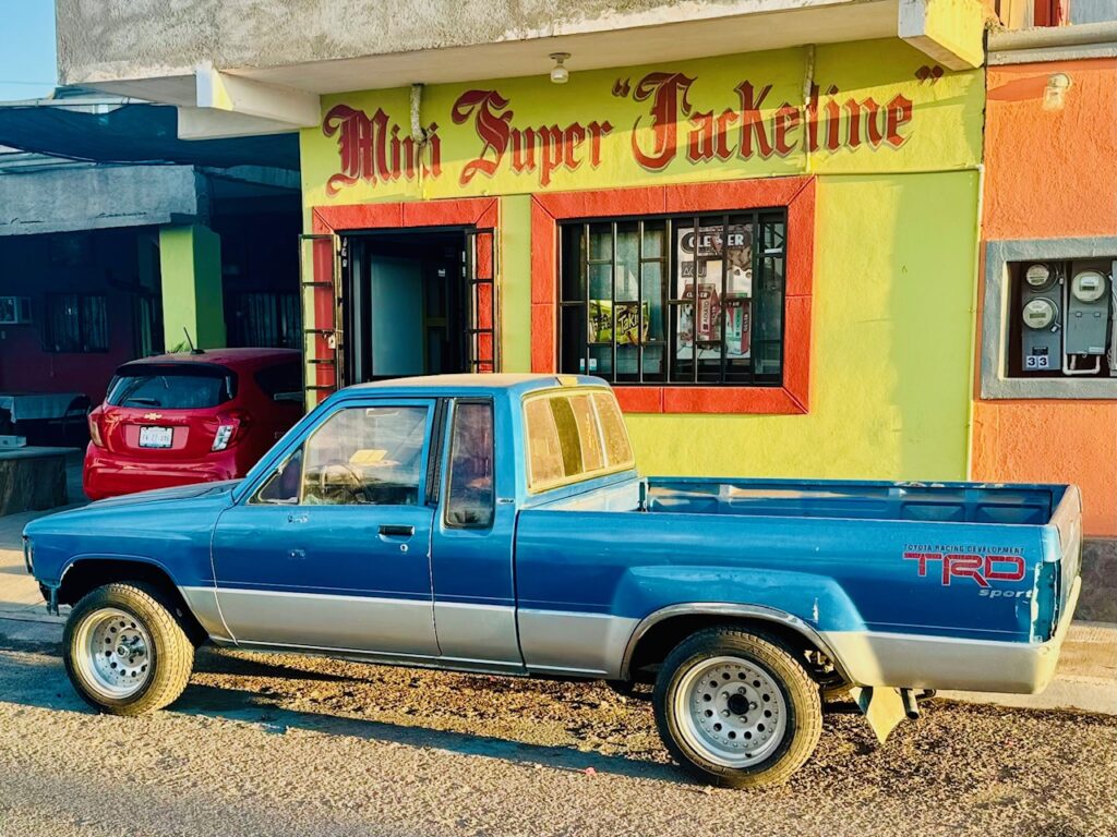 Blauer Pickup mit silberner Zierleiste vor gelb-orangefarbenem Gebäude mit rotem Schriftzug 'Mini Super Jackeline'