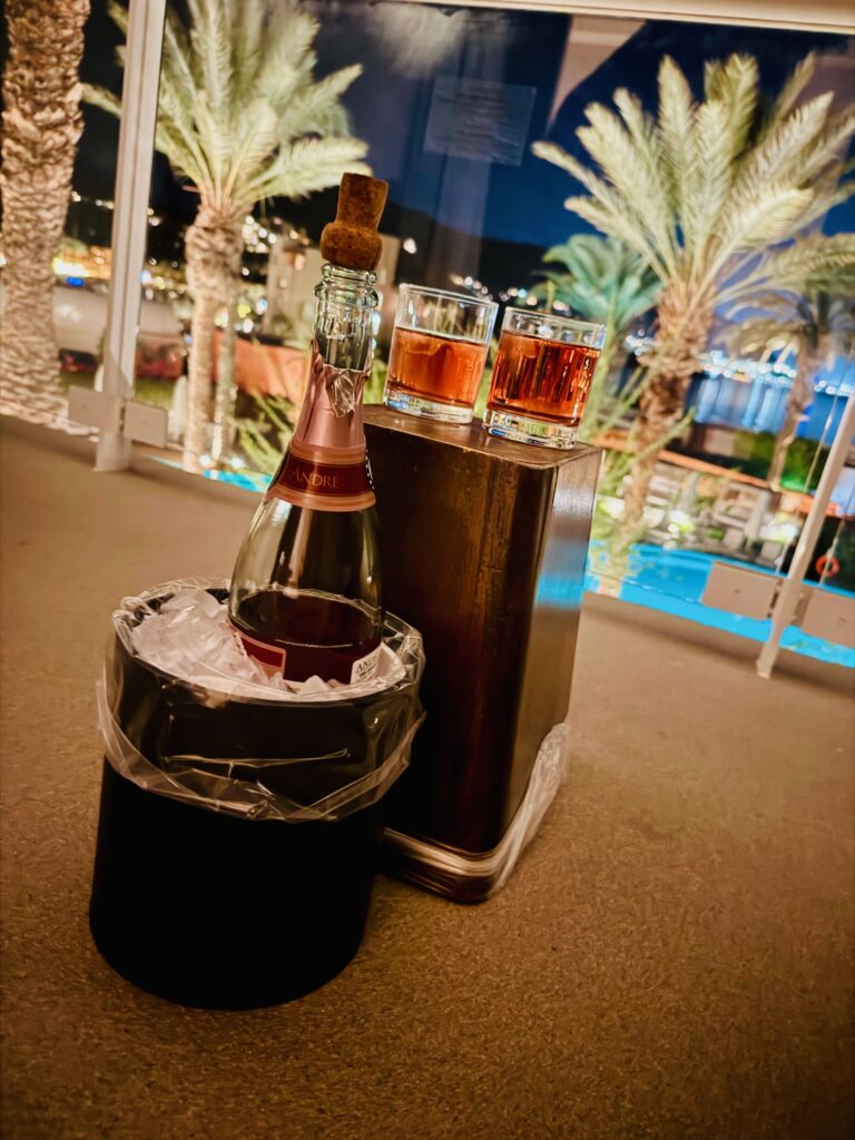 Flasche Sekt in einem Eiskübel, daneben zwei Gläser mit bernsteinfarbenem Getränk, im Hintergrund Palmen und ein beleuchteter Pool