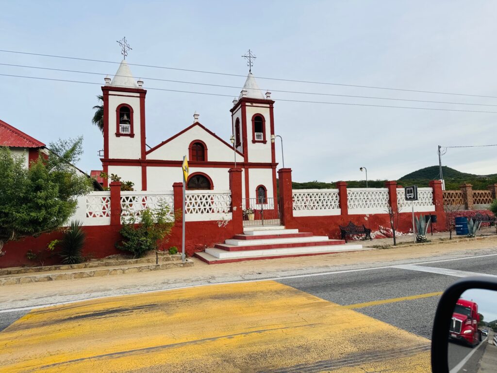 Kirche mit zwei Türmen, weiß mit roten Umrandungen, davor gelb markierter Zebrastreifen und Straße. Im Vordergrund Rückspiegel eines Autos, darin ein Feuerwehrauto