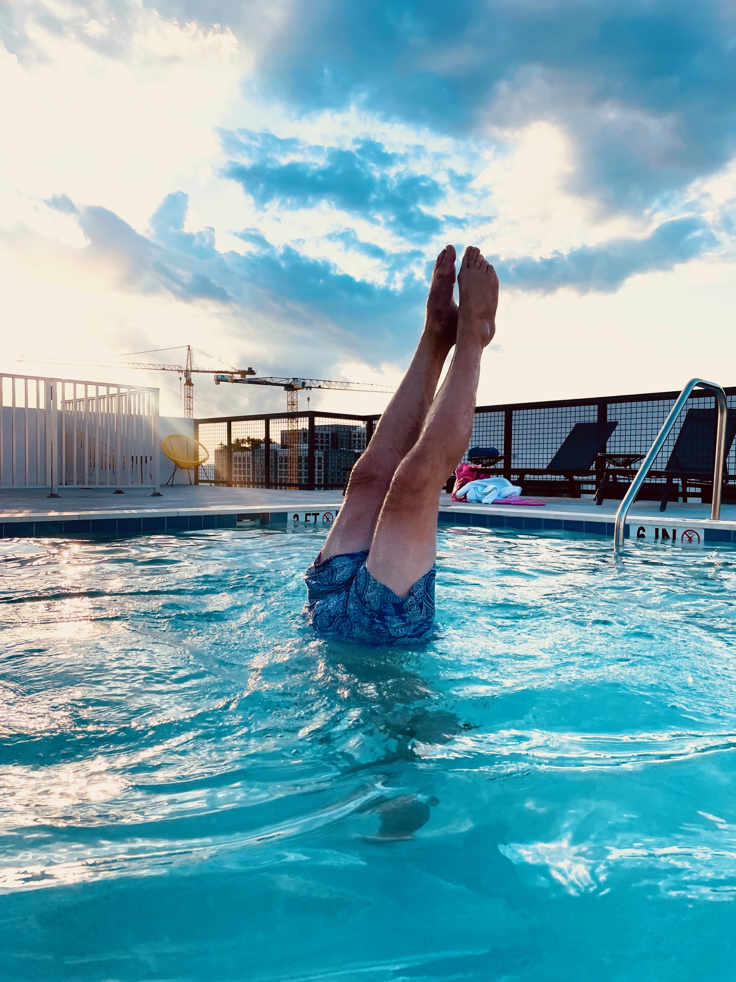 Person in blauer Badehose macht Handstand im Schwimmbecken, nur elegant gestreckte Beine und Füße sichtbar, blauer Himmel mit Wolken im Hintergrund.