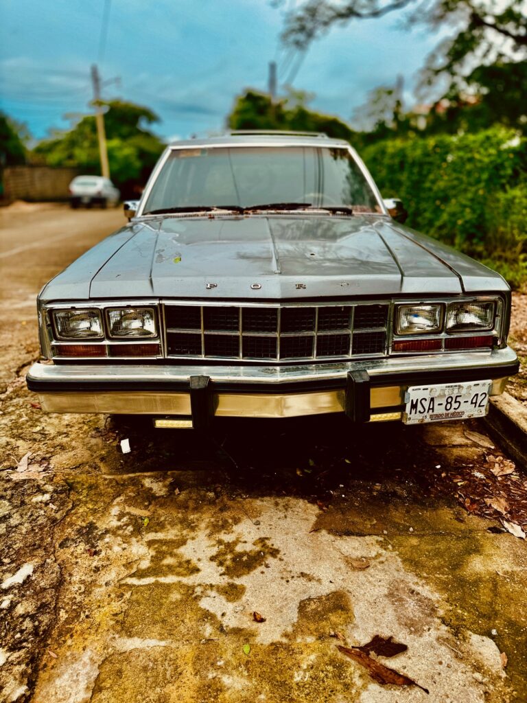 Vorderansicht eines grauen Oldtimers mit rechteckigen Scheinwerfern und mexikanischem Kennzeichen auf einer staubigen Straße mit Bäumen im Hintergrund