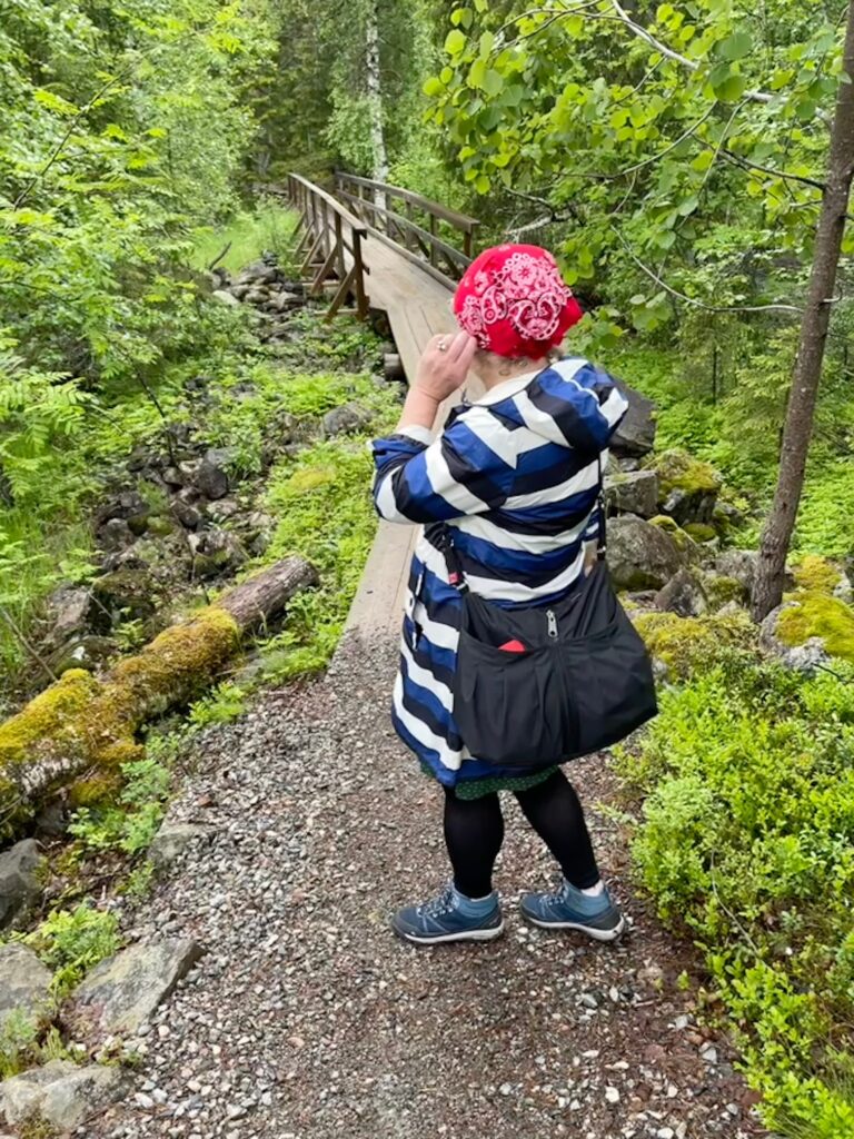 Frau mit rot-weiß gemustertem Kopftuch und blau-weiß gestreiftem Regenmantel steht auf Waldweg vor Holzbrücke, umgeben von grünem, verregneten Laub