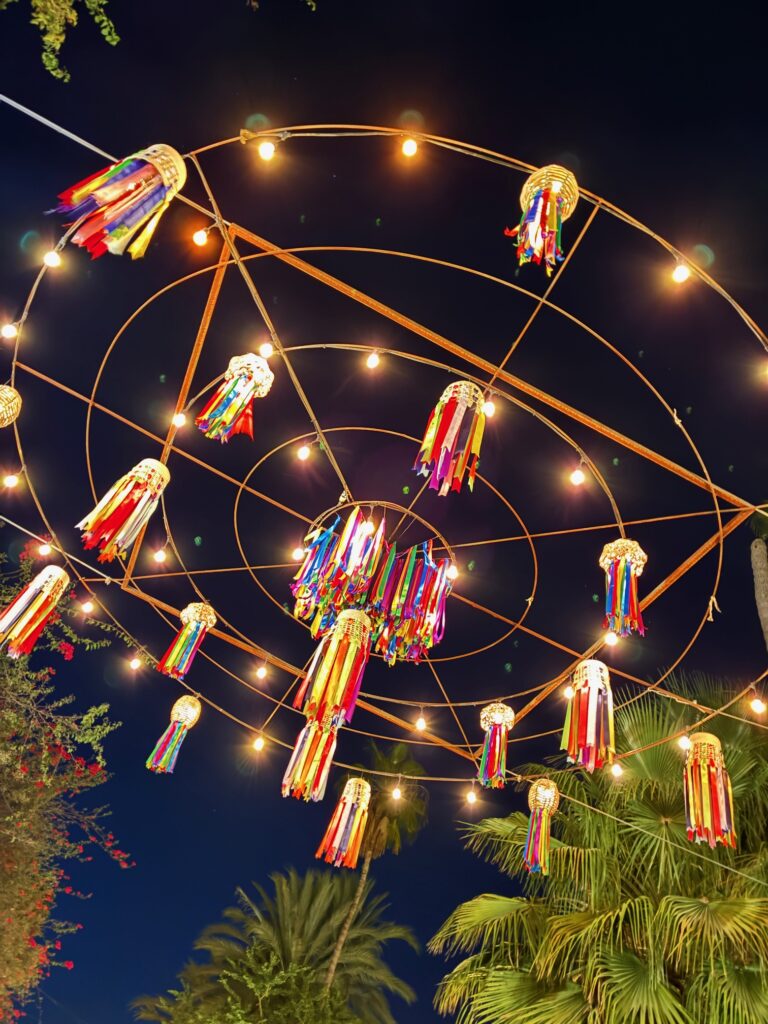 Bunte Papierlaternen mit langen bunten Bändern hängen an einem runden Metallgestell mit Lichterkette vor Nachthimmel und Palmen