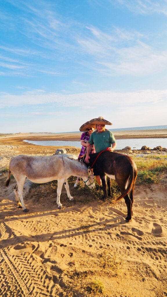 Ein Mann und eine Frau in breitkrempigen Hüten stehen auf einem sandigen Weg an einem Gewässer, zwei Esel fressen am Boden