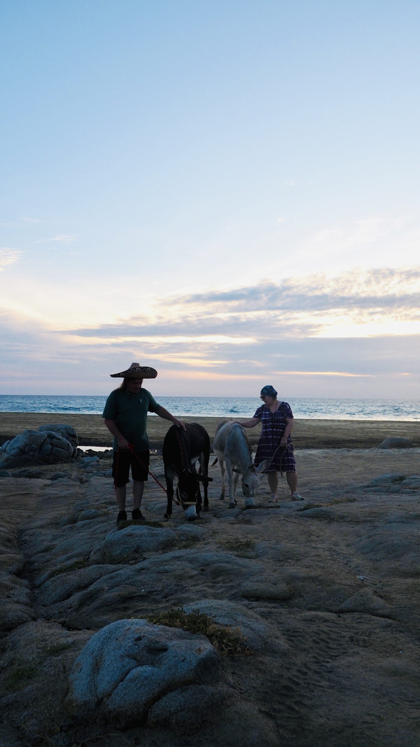 Ein Mann mit Sombrero und eine Frau stehen bei Dämmerung an einer felsigen Küste, zwischen ihnen zwei Esel