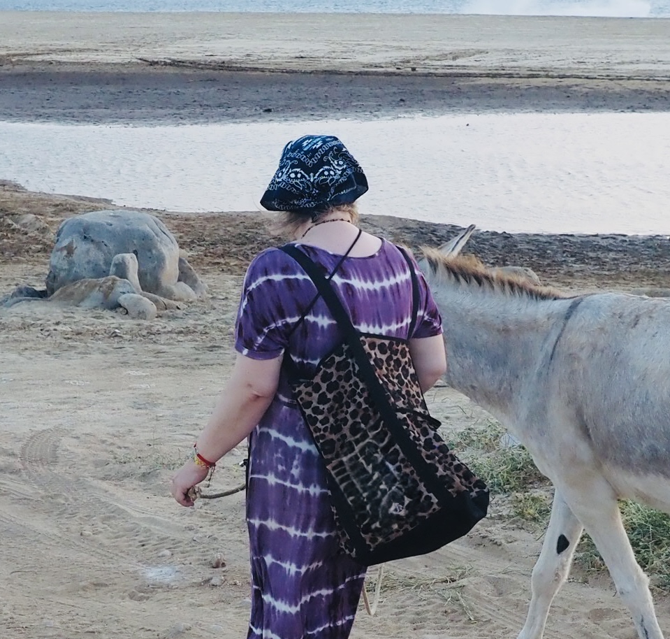 Frau mit blauem Kopftuch und lila Batikkleid geht am Strand neben einem Esel entlang