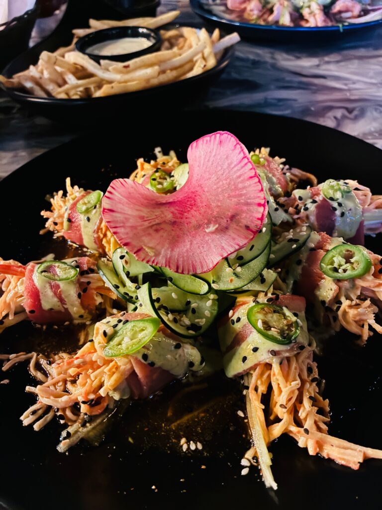 Schwarzer Teller mit mehreren Sushi-Rollen, garniert mit dünnen Gurkenscheiben, Sesam und einer großen Radieschenscheibe in der Mitte