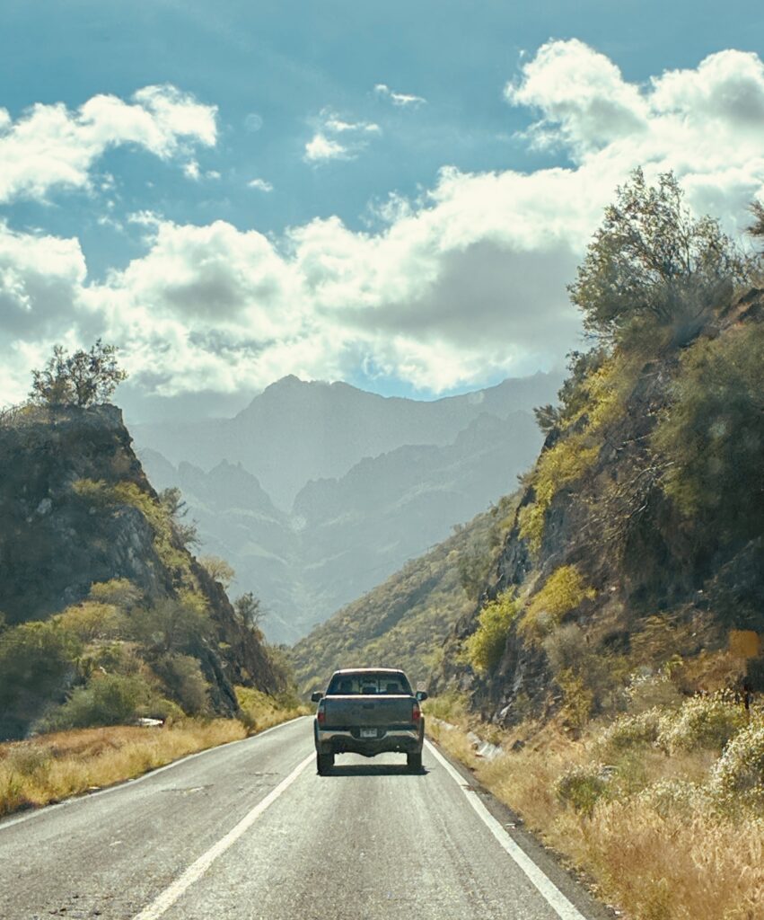 Pick-up fährt auf einer Bergstraße zwischen bewachsenen Hängen unter Himmel mit einigen Wolken