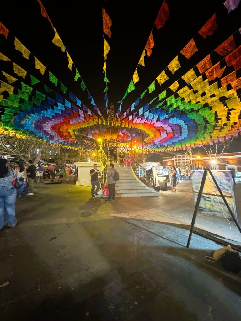 Bunte Papiergirlanden in konzentrischen Kreisen über einem belebten Platz bei Nacht, Menschen stehen und gehen