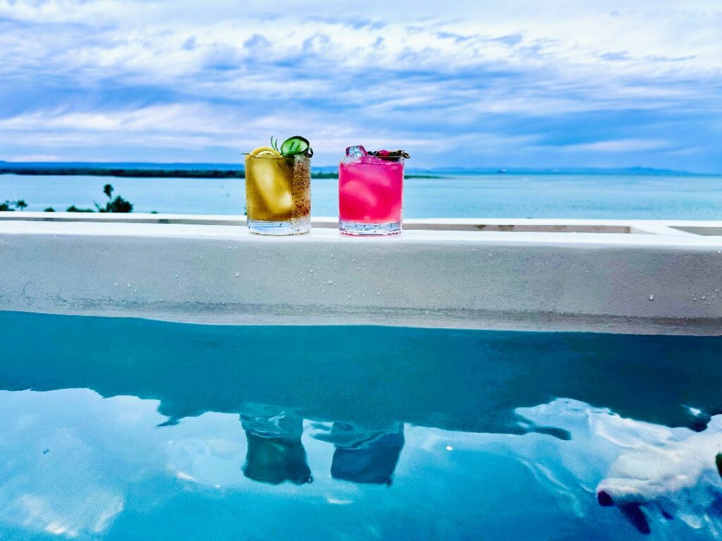 Zwei bunte Cocktails mit Eiswürfeln und Garnitur stehen auf einem weißen Poolrand mit Meer und Himmel im Hintergrund