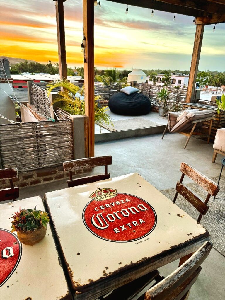 Dachterrasse mit rustikalem Holztisch und Stühlen, Tischplatte mit rotem "Cerveza Corona Extra"-Logo, im Hintergrund Sitzkissen und Blick auf Häuser und Sonnenuntergang