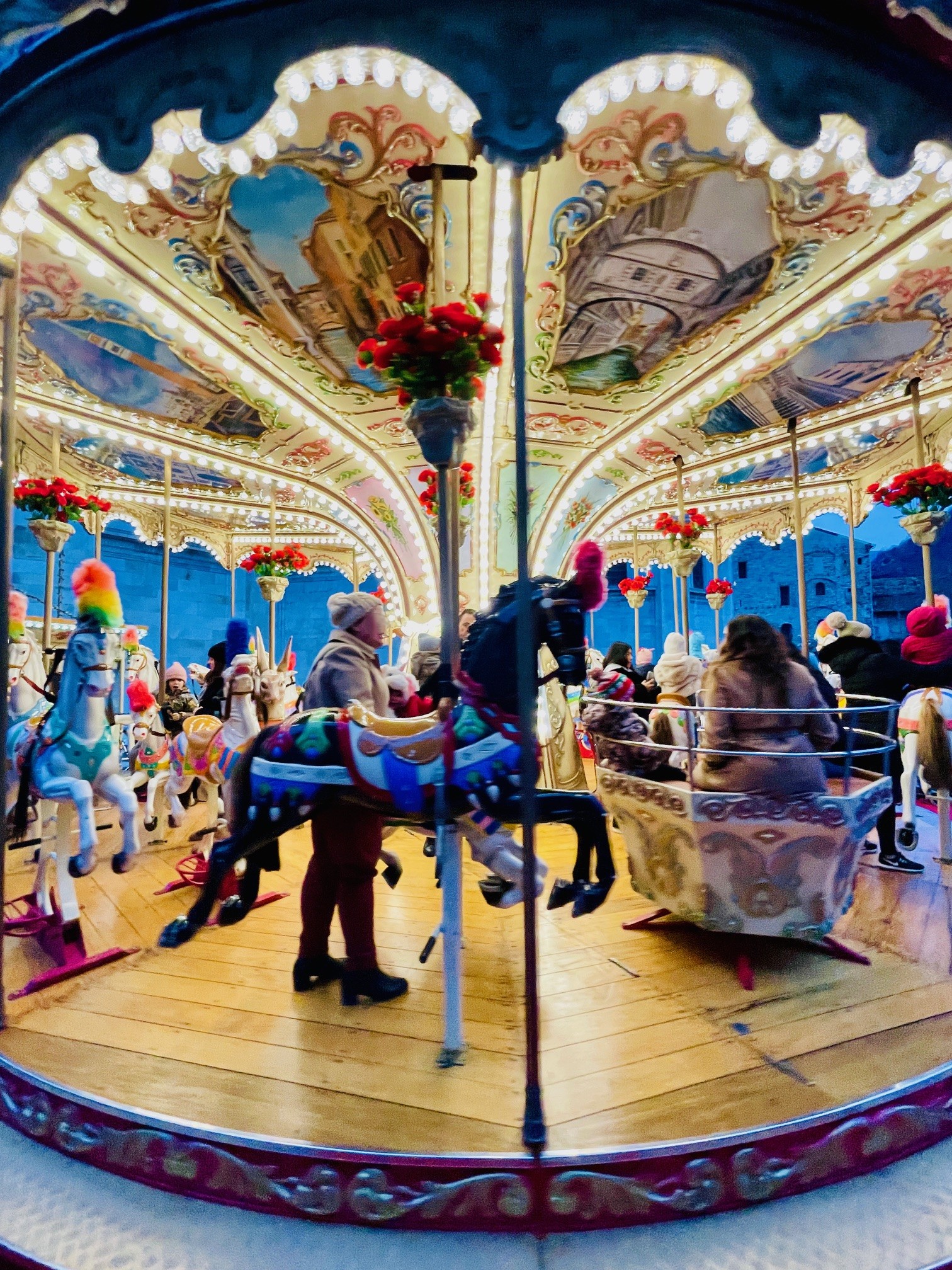 Beleuchtetes Karussell mit bemalten Pferden und Sitzen, Menschen sitzen und stehen darauf, umgeben von Blumenarrangements und dekorativen Deckenmalereien