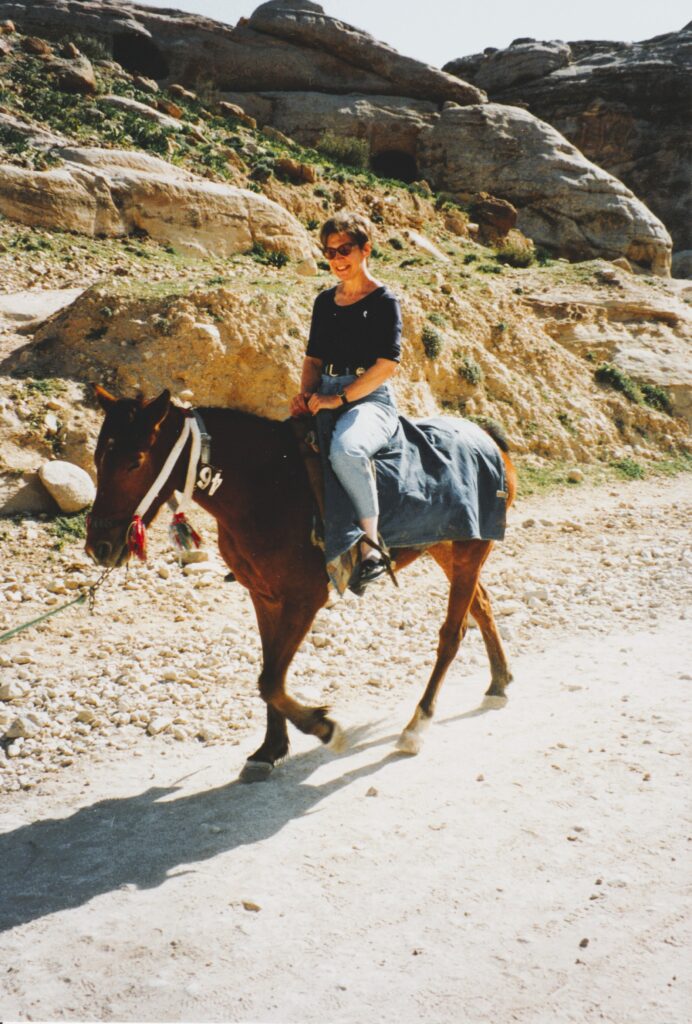 Lächelnde Frau in schwarzem Oberteil und blauer Hose reitet auf braunem Pferd auf einem staubigen Weg vor felsiger Landschaft