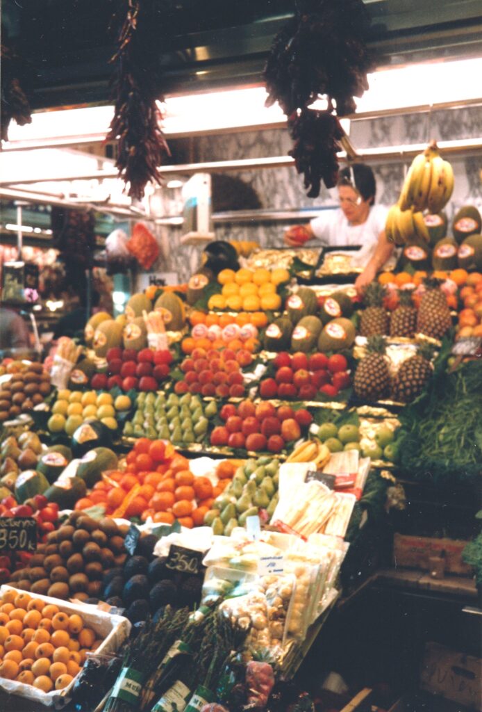 Marktstand mit pyramidenförmig angeordnetem Obst und Gemüse sowie hängenden Chilischoten und Bananen. Historische Analogaufnahme