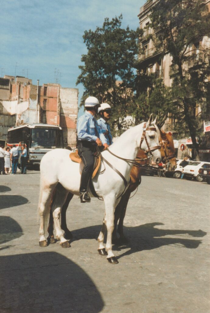 Zwei Polizisten in Uniform und Helm auf weißen Pferden stehen auf einer Straße in einer Stadt mit Gebäuden und parkenden Autos im Hintergrund unter blauem Himmel. Historische Analogaufnahme
