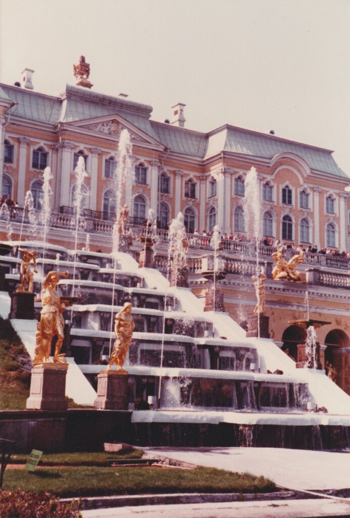 Ausschnitt der Fassade eines barockes Schlosses mit mehrstufigem Springbrunnen und goldenen Statuen im Vordergrund. Es handelt sich um den Peterspalast in St. Petersburg. Historische Analogaufnahme