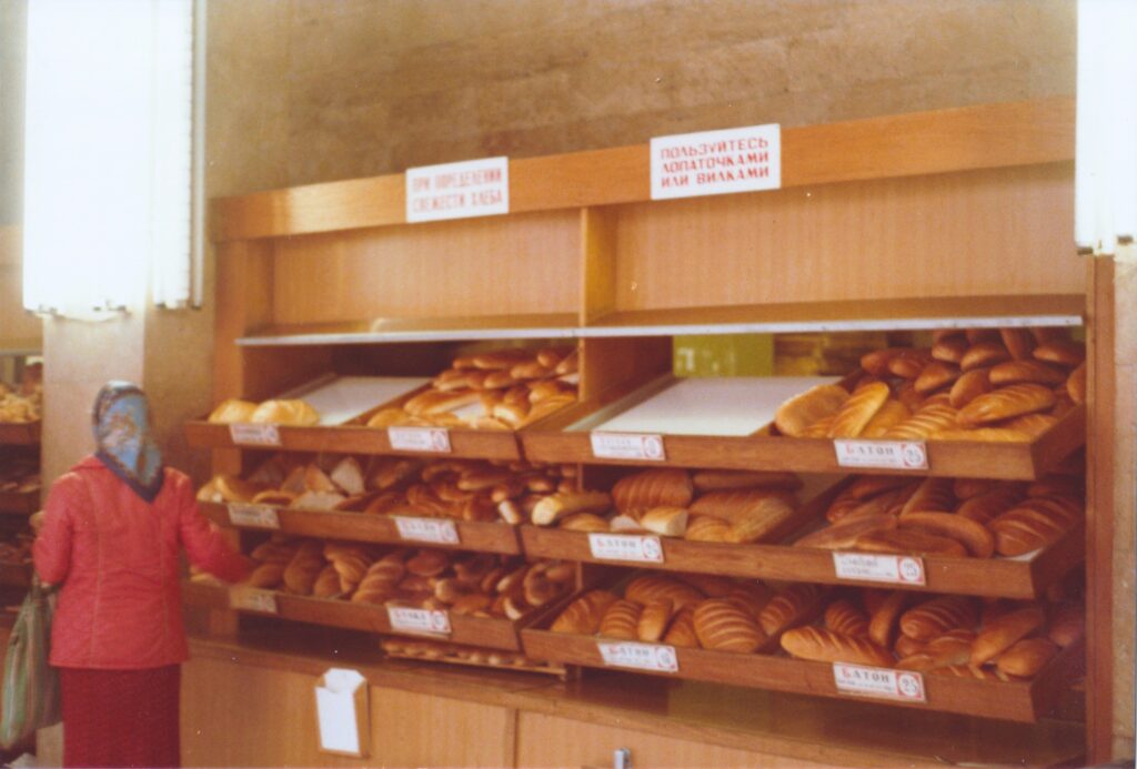 Regal mit verschiedenen Brotsorten in einem Laden, darüber Schilder in kyrillischer Schrift, davor eine Frau mit Kopftuch und rotem Mantel von hinten. Leicht unscharfe historische Analogaufnahme