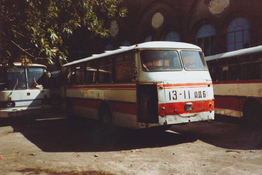 Alter Bus mit weiß-roter, leicht rostiger Lackierung und der Nummer '13-11' steht auf einem Hof vor einem Backsteingebäude. Historische Analogaufnahme