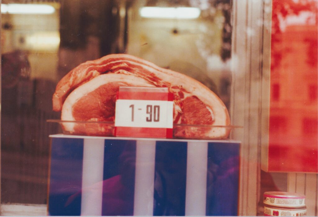 Schaufenster, in dem eine Portion Speck mit einem Preisschild '1-90' auf einem blau-weiß gestreifte Podest liegt, im Hintergrund Neonlampen. Historische Analogaufnahme