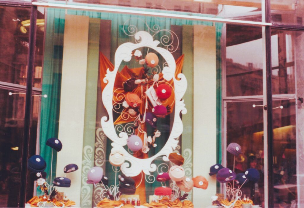 Schaufenster mit bunter Dekoration und vielen auf Ständern präsentierten Hüten in verschiedenen Farben. Leicht unscharfe historische Analogaufnahme