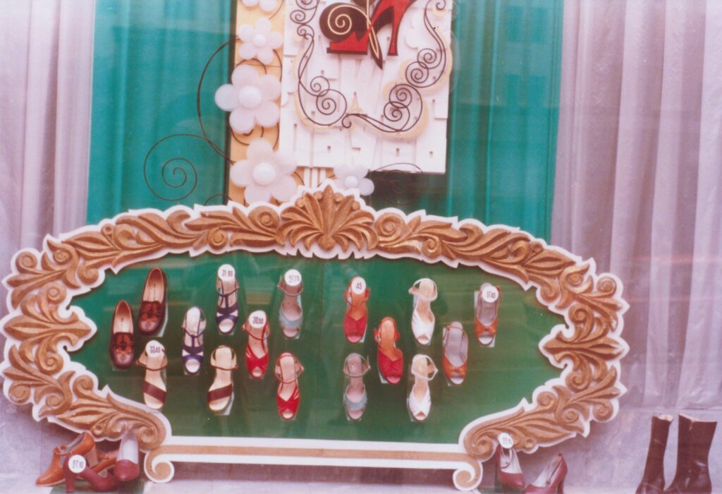 Üppig dekoriertes Schaufenster mit goldverziertem Rahmen, in dem zahlreiche Paar Damenschuhe auf einem grünen Untergrund präsentiert werden. Historische Analogaufnahme