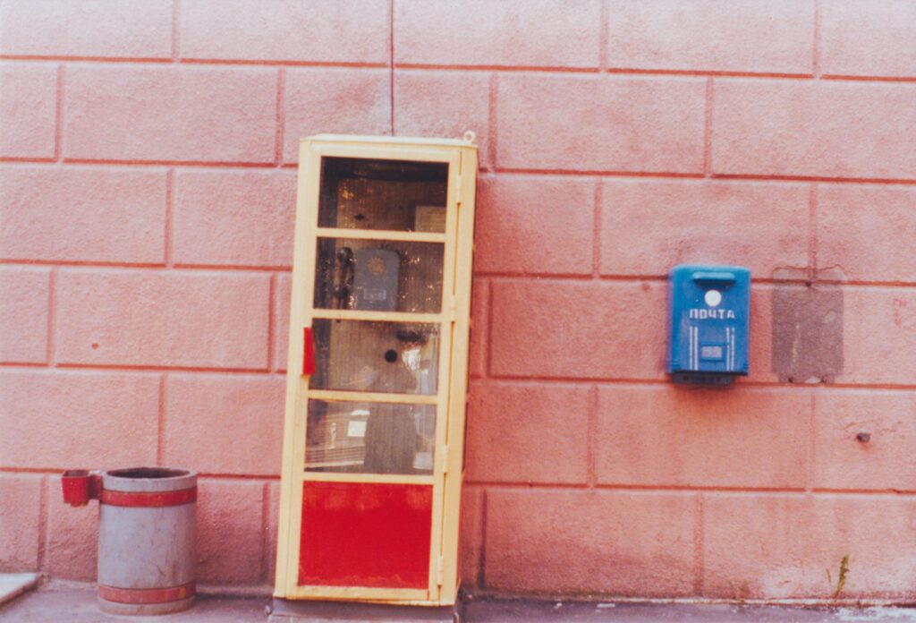 Altmodische, blassgelbe Telefonzelle vor einer rosa Wand neben einem blauen Briefkasten mit kyrillischer Aufschrift und einem grauen Mülleimer. Historische Analogaufnahme