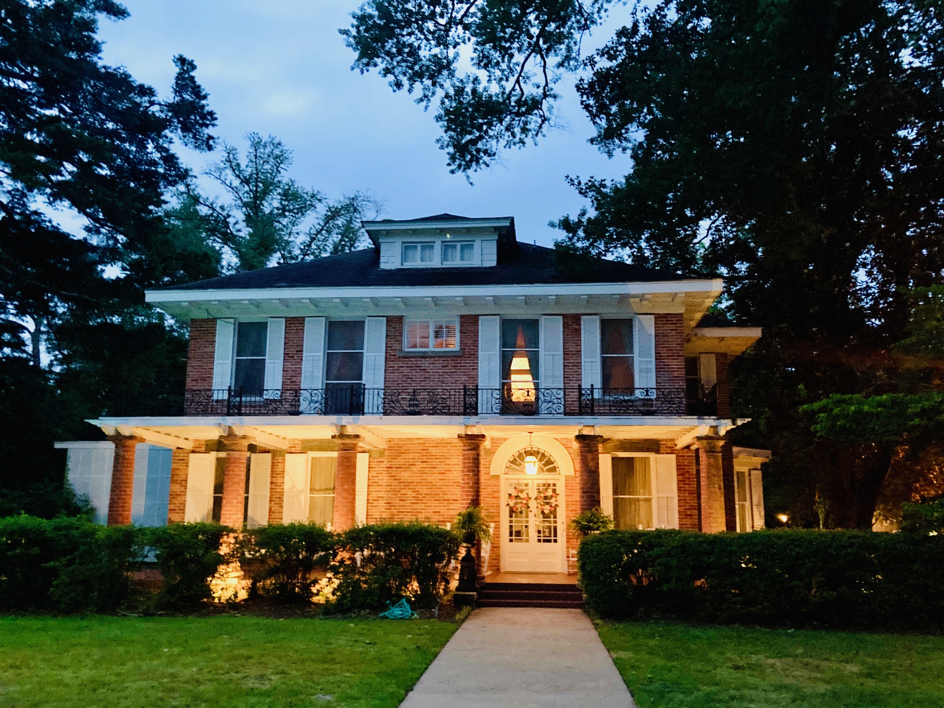 Zweistöckiges Backsteinhaus mit Säulen und großer Rasenfläche davor, dazu stattliche Bäume in der Dämmerung, der untere Teil des Hauses ist beleuchtet