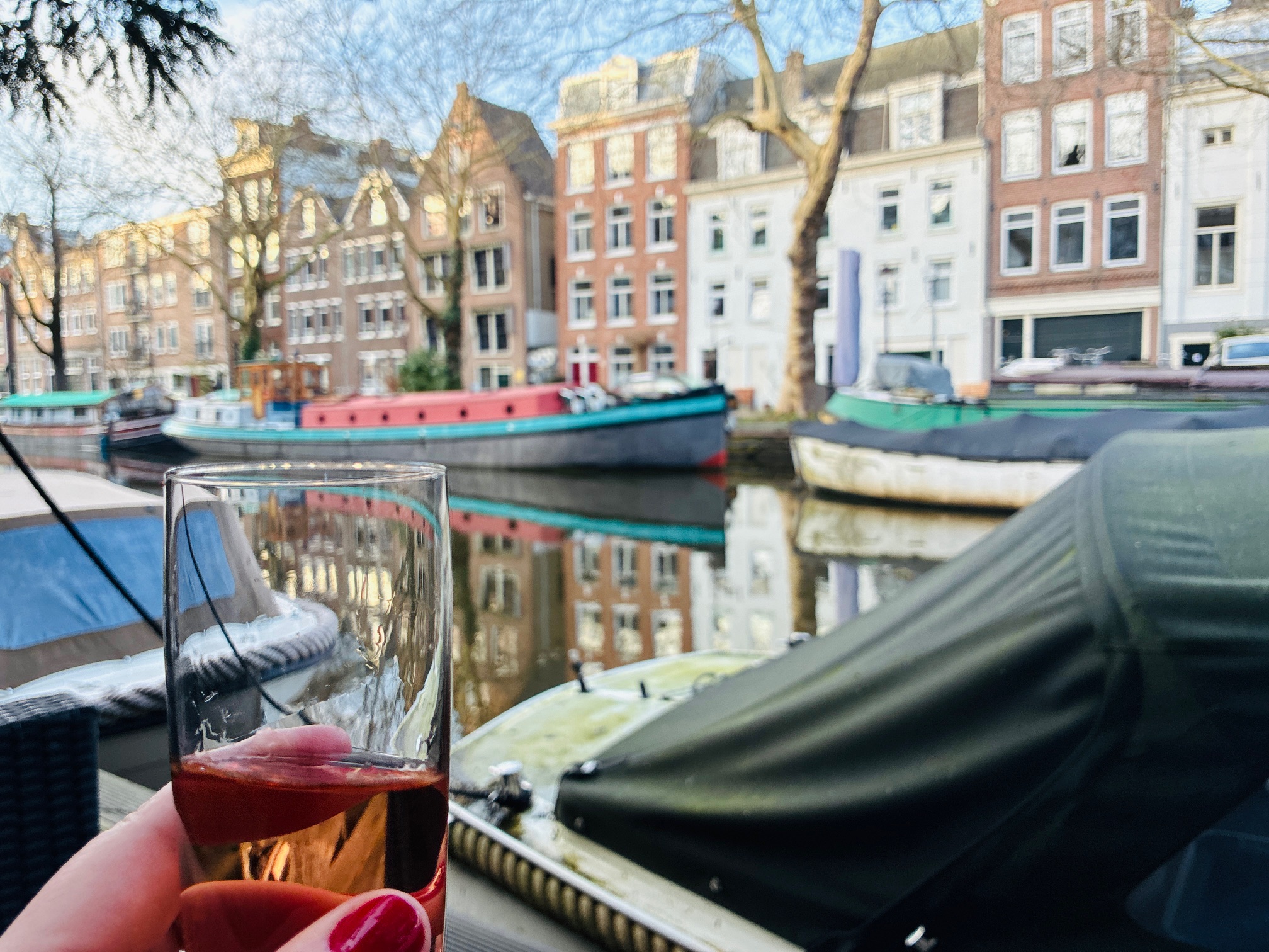 Hand hält ein Glas mit roséfarbenem Getränk vor einem Kanal mit Hausbooten und historischen Giebelhäusern im Hintergrund