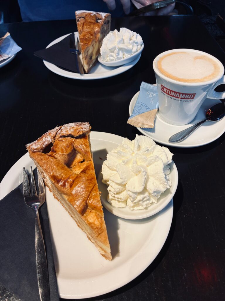 Zwei Teller mit Apfelkuchenstücken und Sahne, dazu eine Tasse Kaffee mit Milchschaum und einem Löffel auf einem schwarzen Tisch