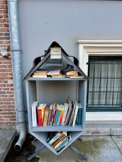 Kleines, graues Bücherregal in Form eines Hauses mit Büchern und Schild 'Gratis Bibliotheek' vor Backsteinwand und Kellerfenster