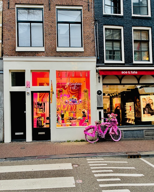 Üppig mit pinken Blumen geschmücktes Fahrrad vor einem Geschäft mit warm beleuchtetem Schaufenster in einer Häuserzeile mit Backsteinfassaden