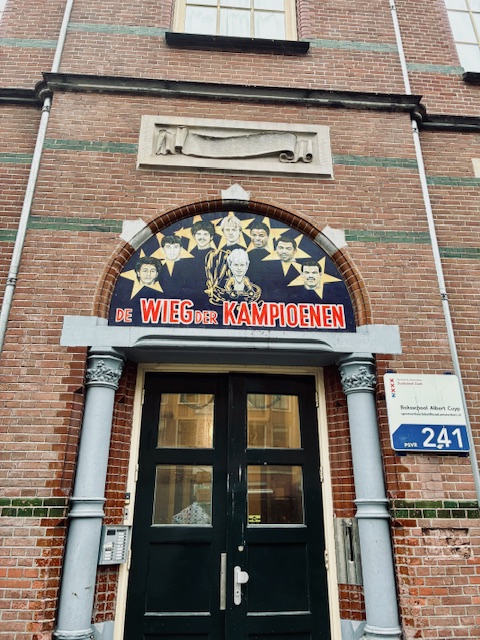 Eingang mit schwarzer Doppeltür in einem Backsteingebäude mit einem halbkreisförmigen Mosaik über der Tür, das mehrere Männerporträts mit Sternen zeigt und den Schriftzug 'DE WIEG DER KAMPIOENEN' enthält