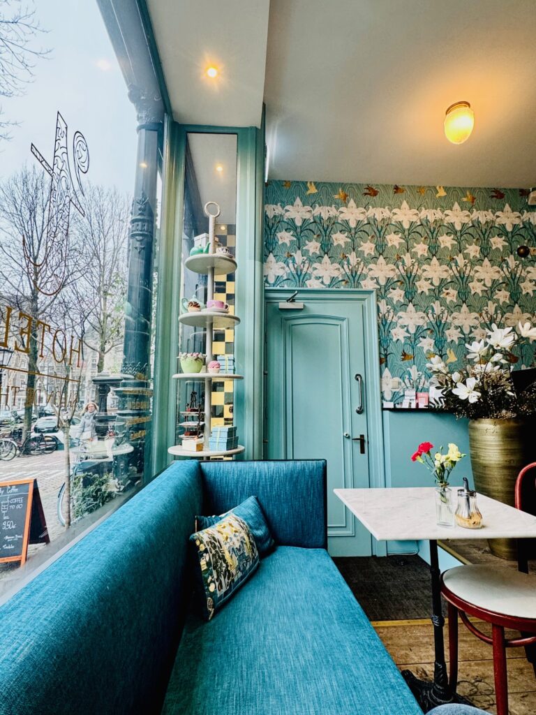 Café-Innenraum mit türkisfarbenem Sitzbanksofa, gemustertem Tapetenhintergrund und kleinem Tisch mit Blumenvasen