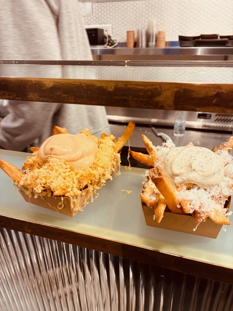 Zwei Pappschalen mit Pommes frites, eine mit Käse und orangener Sauce, die andere mit Käse und weißer Sauce, auf einer Theke