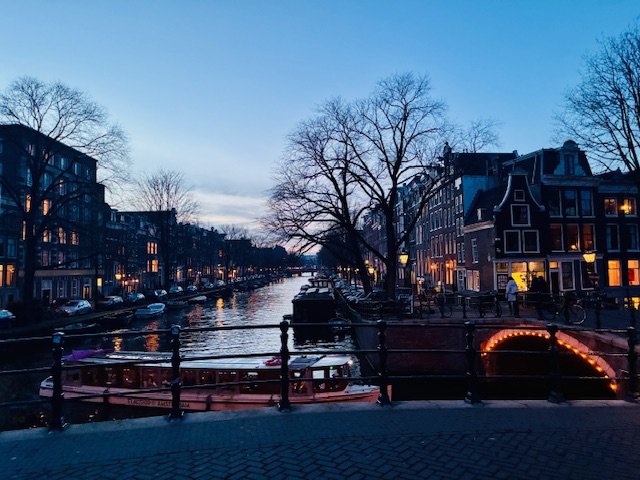 Blick auf einen Kanal in Amsterdam bei Dämmerung mit beleuchteter Brücke, Bäumen ohne Blätter und historischen Häusern