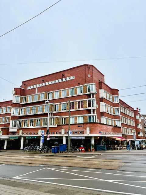Mehrstöckiges rotes Backsteingebäude mit zahlreichen Fenstern und Geschäften im Erdgeschoss an einer Straßenkreuzung mit Straßenbahnschienen