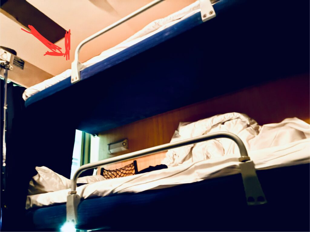 Zwei übereinanderliegende Schlafkojen mit blauen Matratzen und weißen Bettdecken in einem Zugabteil. Auf die obere zeigt ein roter Pfeil