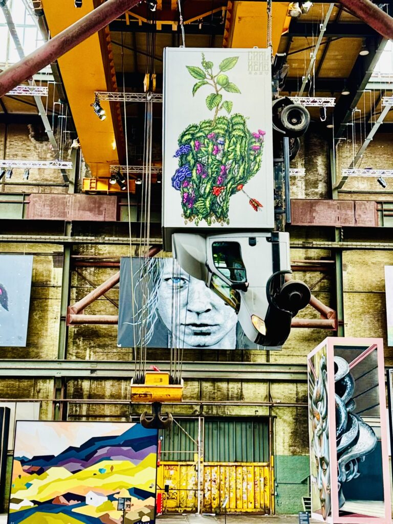 Hängende Banner mit farbigen Kunstwerken und abstrakter Landschaftsmalerei in einer Industriehalle mit sichtbaren Stahlträgern und gelben Toren