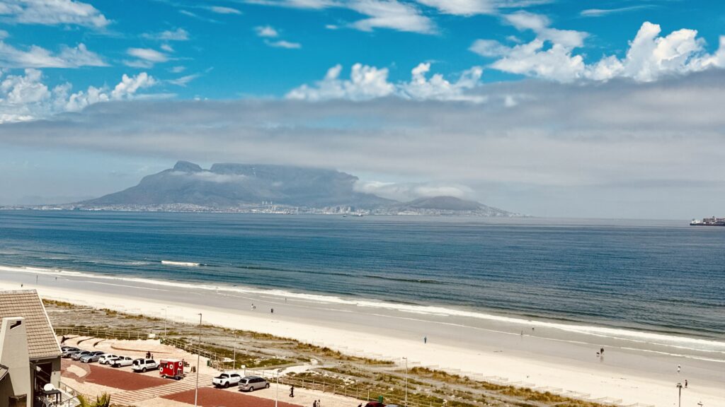 Blick auf einen breiten Sandstrand mit wenigen Menschen und dahinter das Meer sowie der Tafelberg in Kapstadt mit einem Wolkenband unter einem Himmel mit freundlichen Wolken
