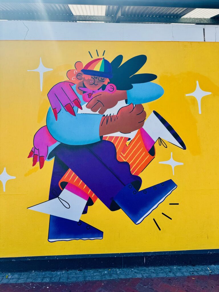 Bunte Wandmalerei zeigt zwei Personen in Umarmung vor gelbem Hintergrund mit weißen Sternsymbolen