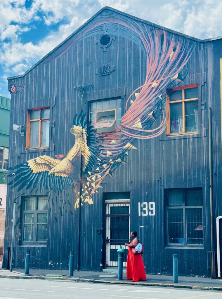 Fassade eines Gebäudes mit großem Wandgemälde eines stilisierten Vogels mit ausgebreiteten Flügeln und einer Person in rotem Kleid davor