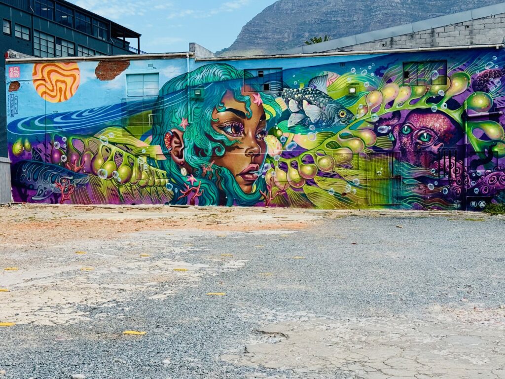 Bunte Wandmalerei mit einem großen Fisch, einer menschlichen Figur und abstrakten, wellenförmigen Mustern vor einem Industriegebäude und Tafelberg in Kapstadt im Hintergrund