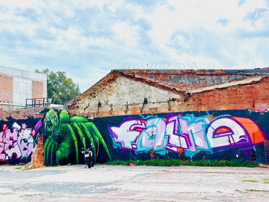 Großflächiges Wandgraffiti mit einer grünen Spinne und buntem Schriftzug auf einer Mauer vor einem alten Backsteingebäude