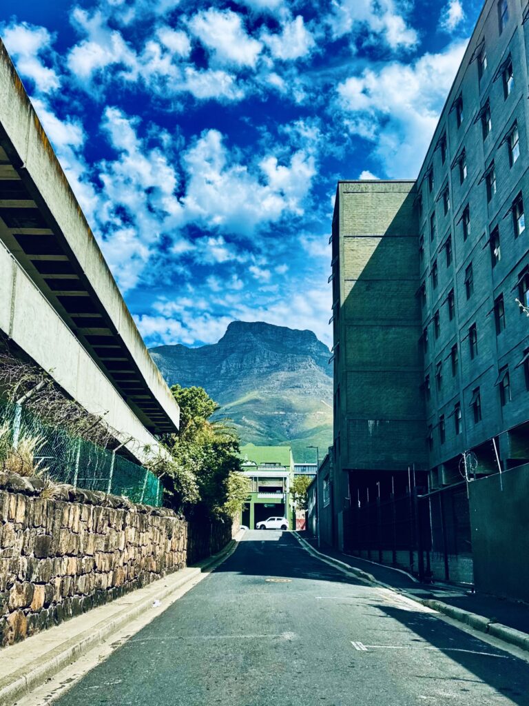 Gasse mit links einer Mauer und rechts einem mehrstöckigen Gebäude vor dem Tafelberg in Kapstadt unter blauem Himmel mit freundlichen Wolken