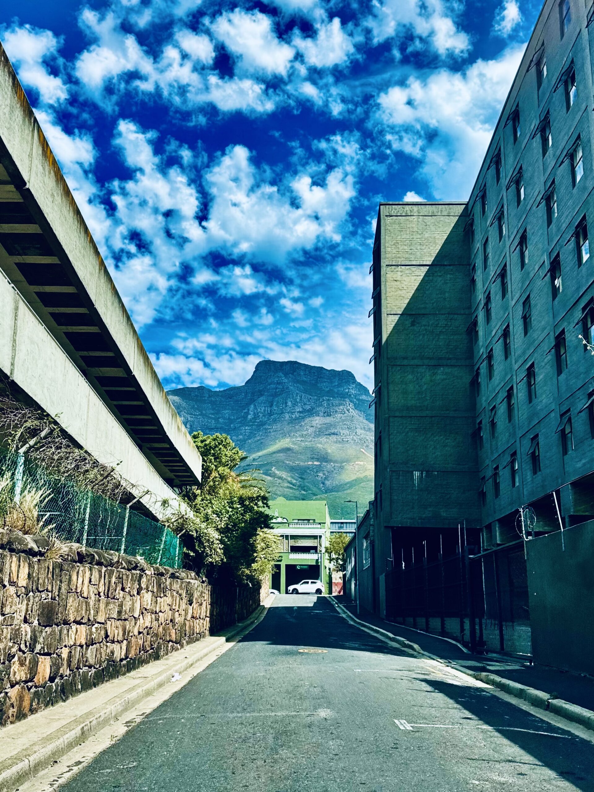 Gasse mit links einer Mauer und rechts einem mehrstöckigen Gebäude vor dem Tafelberg in Kapstadt unter blauem Himmel mit freundlichen Wolken